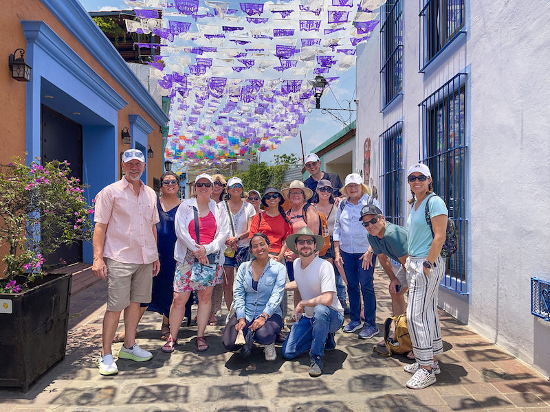 Group of Spanish immersion retreat participants smiling on a colorful street under papel picado in Jalatlaco, Oaxaca, Mexico.